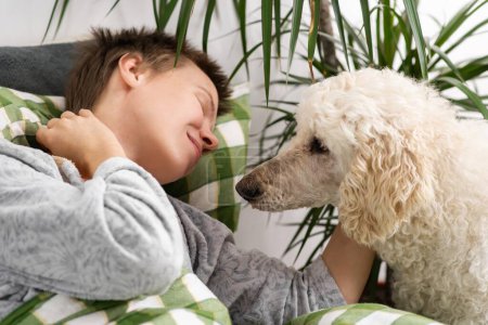 Engrossed in leisure, a middle-aged woman with short hair spends her time in bed with her big poodle. Laziness, weekend relaxation