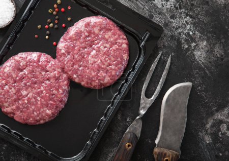 Photo for Raw beef burgers sealed in vacuum tray with barbeque fork and knife on dark kitchen background.Top view. - Royalty Free Image