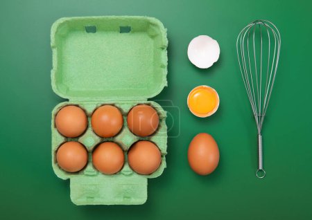 Téléchargez les photos : Raw brown eggs in paper tray with whisk,yolk and shell on green background.Top view - en image libre de droit