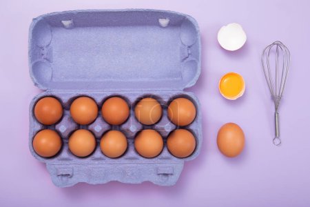 Foto de Tray with organic brown eggs with yolk,whisk and shell on purple.Top view concept - Imagen libre de derechos