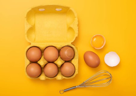 Photo for Raw brown eggs in paper tray with whisk,yolk and shell on yellow background.Top view - Royalty Free Image
