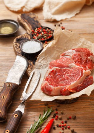 Photo for Rib eye raw fillet  with fork and knife,salt and pepper with rosemary on wooden table with herbs and oil. - Royalty Free Image
