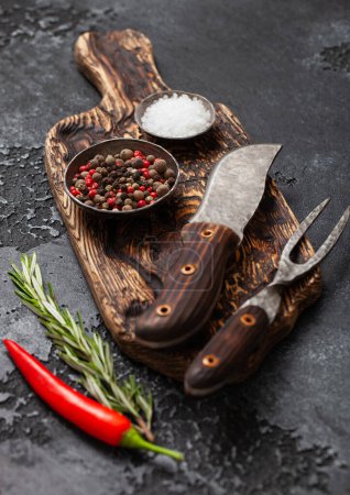 Photo for Fork and knife on chopping board with pepper and salt with rosemary on black kitchen board. - Royalty Free Image