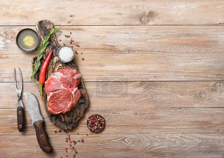 Photo for Rib eye raw fillet  with fork and knife,salt and pepper with rosemary on wooden kitchen table.Top view. - Royalty Free Image