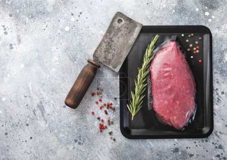 Photo for Raw sirloin ribeye fillet steak in vacuum tray with meat cleaver and rosemary with pepper on light background.Top view. - Royalty Free Image