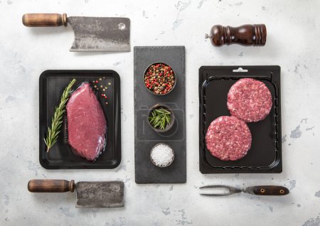 Foto de Raw fresh beef sirloin fillet steak and mince burgers sealed in vacuum tray on light  background with barbeque utensils.Top view. - Imagen libre de derechos
