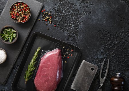 Photo for Raw beef fillet steak in vacuum tray with pepper on on black background with meat cleaver and fork. Top view. - Royalty Free Image