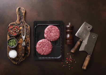Foto de Raw beef burgers sealed in vacuum tray with cleavers on dark kitchen table with pepper grinder and salt..Top view. - Imagen libre de derechos