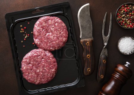 Photo for Raw beef burgers sealed in vacuum tray with barbeque fork and knife on dark background with pepper grinder. - Royalty Free Image
