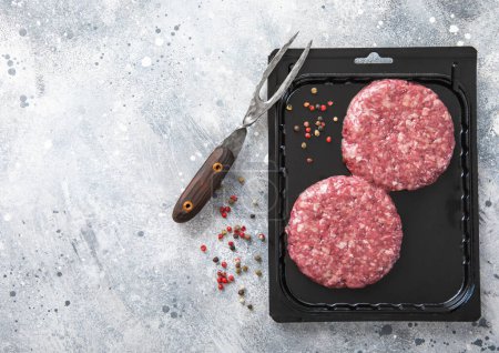 Foto de Beef fresh raw burgers in vacuum tray with fork and pepper on light background.Top view. - Imagen libre de derechos
