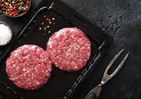 Photo for Beef organic burgers in vacuum tray with fork and salt and pepper on black. - Royalty Free Image