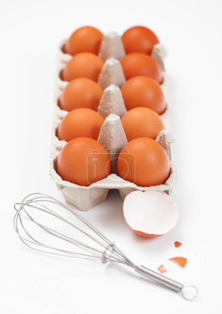 Foto de Raw brown organic eggs in paper tray on white with whisk and shell - Imagen libre de derechos