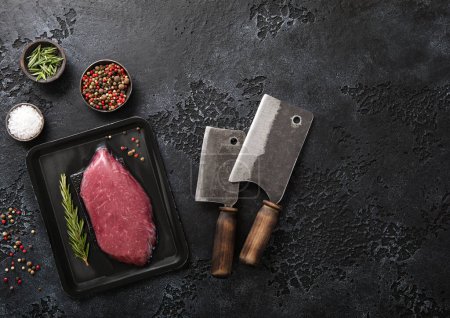 Foto de Beef organic raw fillet steak in vacuum tray with meat cleavers on dark background with rosemary ,salt and pepper. Top view. - Imagen libre de derechos