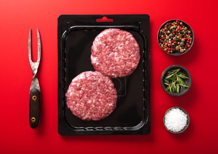 Foto de Beef burgers in vacuum tray with fork and salt and pepper on red background. Top view - Imagen libre de derechos