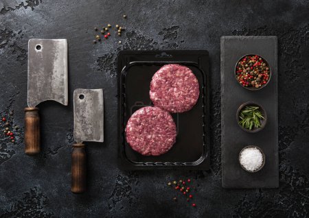 Photo for Raw beef burgers sealed in vacuum tray with cleavers and salt and pepper on black background. Top view - Royalty Free Image