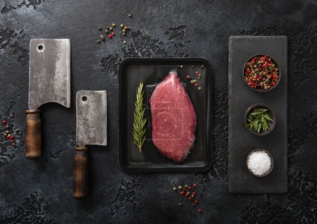 Photo for Raw beef fillet steak in vacuum tray with pepper,salt and rosemary on on black background with meat cleavers.Top view. - Royalty Free Image
