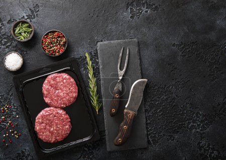 Téléchargez les photos : Raw beef burgers sealed in vacuum tray with barbeque fork and knife on dark kitchen background with spices. - en image libre de droit