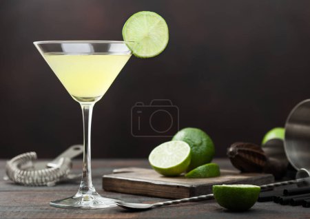 Foto de Cóctel Gimlet Kamikaze en vaso de martini con rodaja de lima y hielo sobre fondo de madera con limas frescas y colador con coctelera. - Imagen libre de derechos
