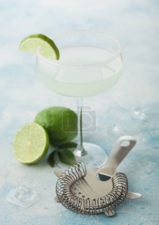 Foto de Crystal glass of Margarita cocktail with fresh limes and strainer on light blue table background. Macro - Imagen libre de derechos