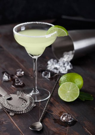 Foto de Classic glass of Margarita cocktail with fresh limes and bar spoon with strainer and ice cubes on wooden table background. Popular summer cocktail - Imagen libre de derechos