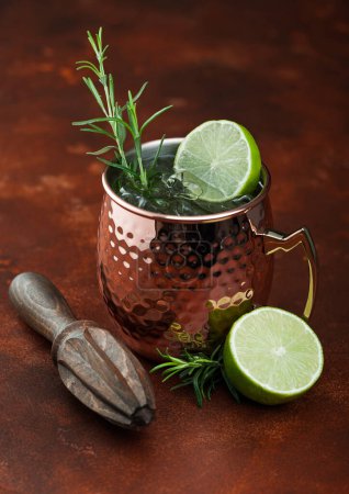 Photo for Moscow mule cocktail in a copper mug with lime and rosemary and wooden squeezer on brown kitchen table background - Royalty Free Image