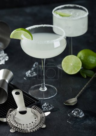 Photo for Luxury glasses of Margarita cocktail with fresh limes and bar mat with strainer and jigger, with spoon and ice cubes on black table background. - Royalty Free Image
