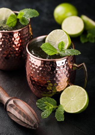 Photo for Moscow mule cocktail in a copper mug with lime and mint and wooden squeezer on dark kitchen background. - Royalty Free Image