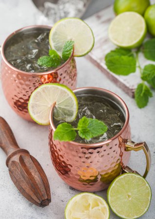 Photo for Moscow mule cocktail in a copper mug with lime and mint and wooden squeezer on light kitchen table background with steel shaker - Royalty Free Image