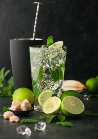 Photo for Glass of Mojito cocktail with ice cubes mint and lime on black board with spoon in shaker and wooden squeezer and fresh limes. Summer party drink. - Royalty Free Image