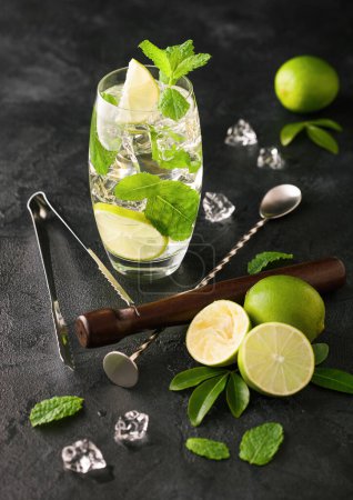 Téléchargez les photos : Verre de cocktail Mojito avec glaçons menthe et citron vert sur panneau noir avec cuillère et fouetteur et limes fraîches
. - en image libre de droit
