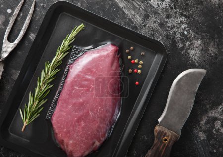 Photo for Beef raw fillet steak in vacuum tray with barbeque fork and knife on black background with spices.Top view. - Royalty Free Image