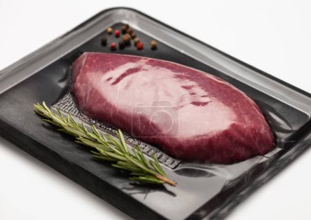 Téléchargez les photos : Steak de filet de boeuf cru frais scellé dans un plateau à vide avec poivre et romarin sur blanc. - en image libre de droit