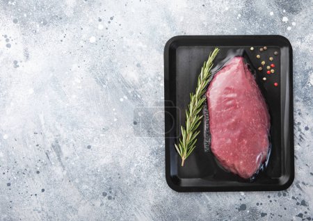 Photo for Fresh raw beef fillet steak sealed in vacuum tray with pepper and rosemary on light kitchen table background. Space for text. - Royalty Free Image