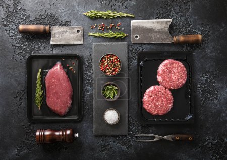 Photo for Fresh raw beef fillet steak and mince burgers in vacuum tray with cleavers and rosemary on black background with rosemary.Top view. - Royalty Free Image