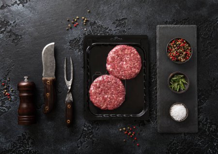 Photo for Raw minced beef burgers sealed in tray on black stone background with fork and knife.Top view. - Royalty Free Image