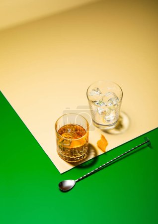 Foto de Vidrio de whisky con cuchara de bar y vidrio vacío con cubitos de hielo sobre fondo dorado gre con cáscara de naranja - Imagen libre de derechos