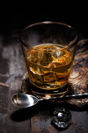 Photo for Glass of single malt whiskey with bar spoon and ice cubes on wooden background.Macro. - Royalty Free Image