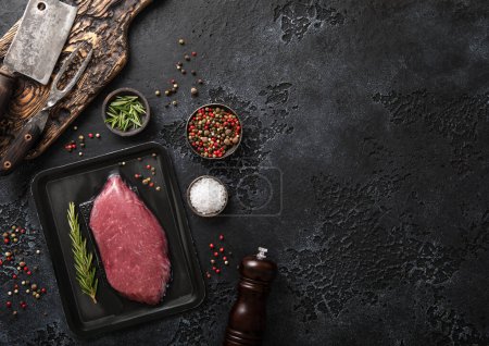 Photo for Raw beef fillet steak in vacuum tray with rosemary on on black background with meat cleavers and pepper grinder.Top view. - Royalty Free Image