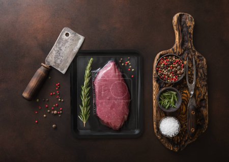 Photo for Chopping board with salt,pepper and rosemary with raw beef fillet sirloin steak in sealed tray on dark background with meat cleaver.Top view. - Royalty Free Image