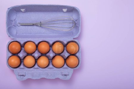 Photo for Brown raw eggs in paper tray with whisk on purrple background.Top view - Royalty Free Image