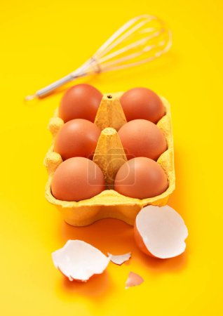 Photo for Brown raw eggs in paper tray with whisk,yolk and shell on yellow. - Royalty Free Image