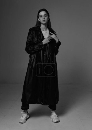 Téléchargez les photos : Jeune beau mannequin en long manteau en cuir et jean foncé sur gris foncé. - en image libre de droit