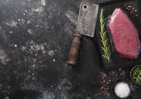 Photo for Raw sirloin ribeye fillet steak in vacuum tray with meat cleaver and rosemary with pepper on dark background.Top view. - Royalty Free Image