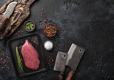 Photo for Raw beef fillet steak in vacuum tray with rosemary on on black background with meat cleavers and barbeque fork and knife.Top view. - Royalty Free Image