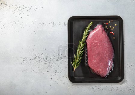 Téléchargez les photos : Steak de filet de bœuf cru frais scellé dans un plateau sous vide avec poivre et romarin sur fond de table de cuisine légère. Vue du dessus. - en image libre de droit