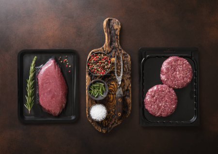 Téléchargez les photos : Steak de filet de bœuf frais cru et hamburgers hachés scellés dans un plateau à vide sur fond sombre avec des épices et une fourchette. - en image libre de droit