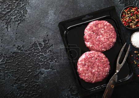 Foto de Hamburguesas de carne de res orgánica cruda selladas en bandeja de vacío con tenedor y pimienta en el fondo de la cocina oscura.. - Imagen libre de derechos