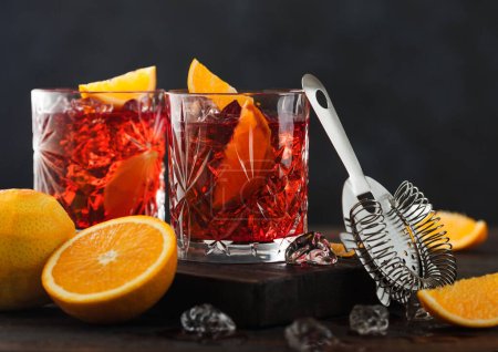 Photo for Negroni cocktail in crystal glasses with orange slice and fresh raw oranges on chopping board with strainer on wooden background. - Royalty Free Image