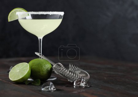 Glass of Margarita cocktail with fresh limes and strainer with ice cubes on wooden table background. Space for text