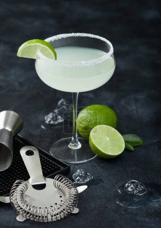 Téléchargez les photos : Verre de luxe de cocktail Margarita avec limes fraîches et tapis de bar avec passoire et jigger sur fond de table noire. - en image libre de droit